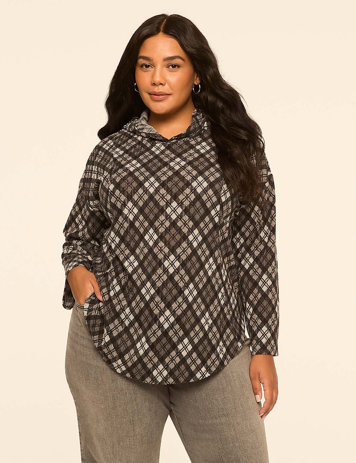 A woman with long, dark, wavy hair stands in the foreground wearing a gray and black plaid hoodie and gray jeans, against a plain, light beige background. Product Image 1