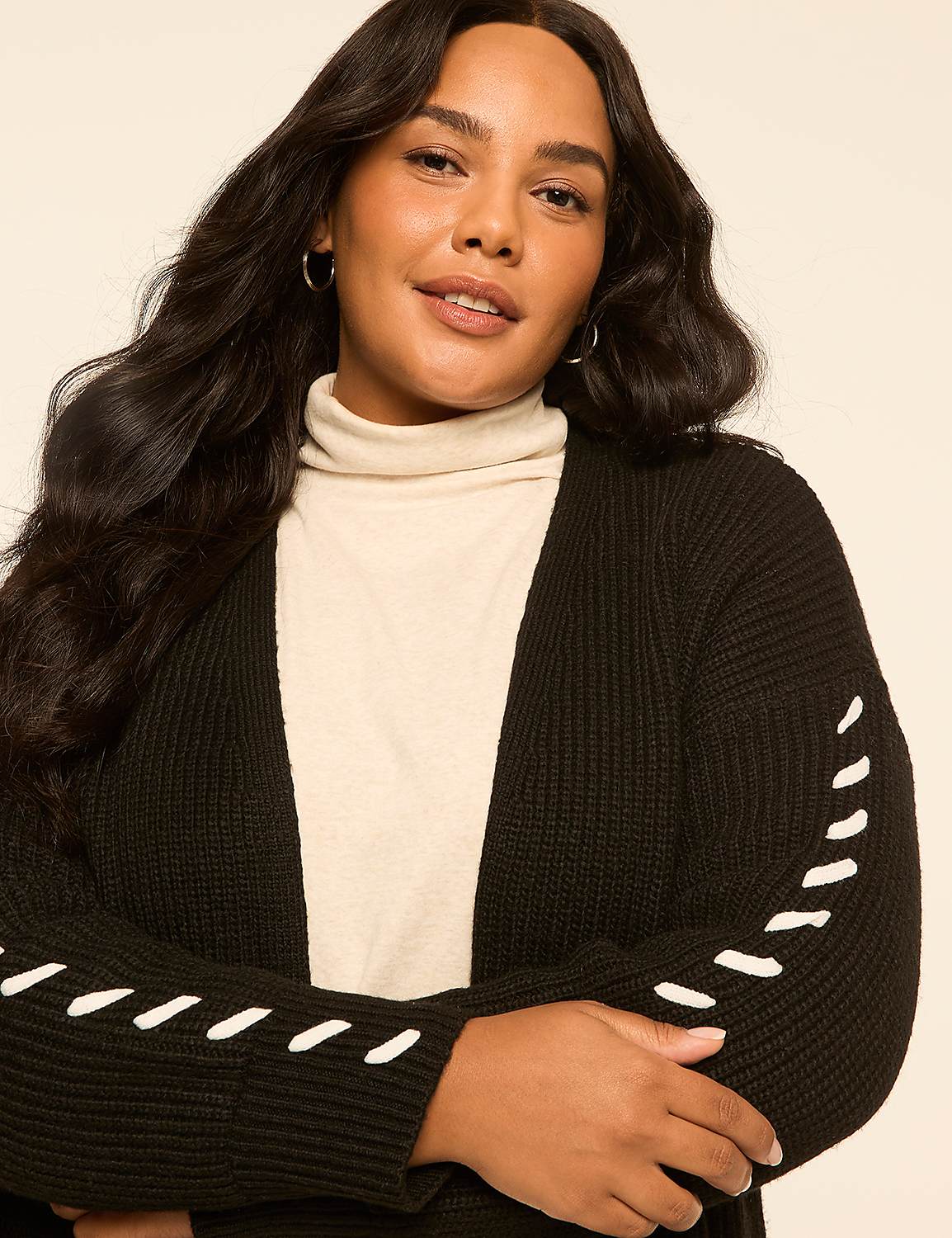 A woman with dark, wavy hair and a light complexion smiles gently, wearing a cream turtleneck and a black cardigan with white stitching on the sleeves, against a plain, light beige background. Product Image 4