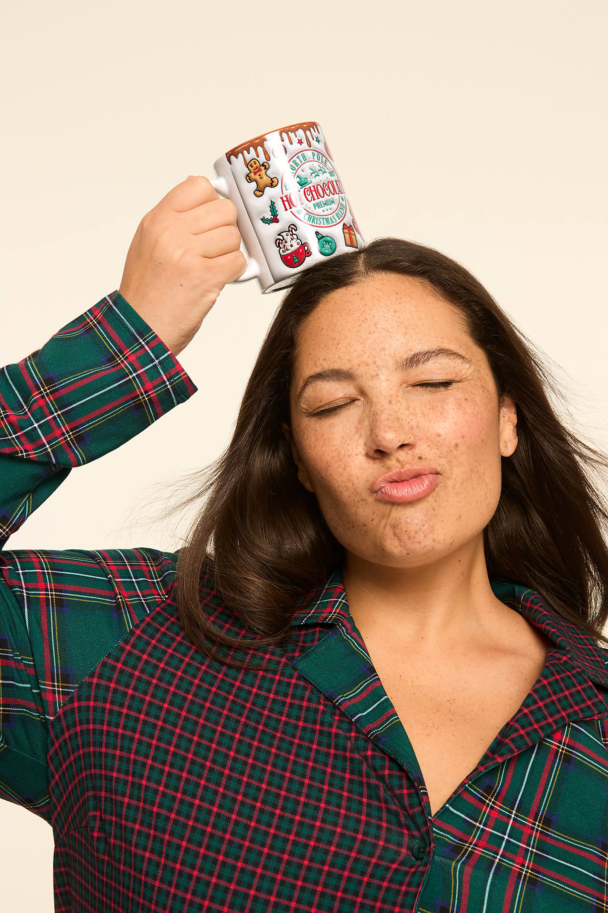A woman in a plaid pajama top balances a Christmas-themed mug on her head while puckering her lips against a plain, light background. Product Image 10