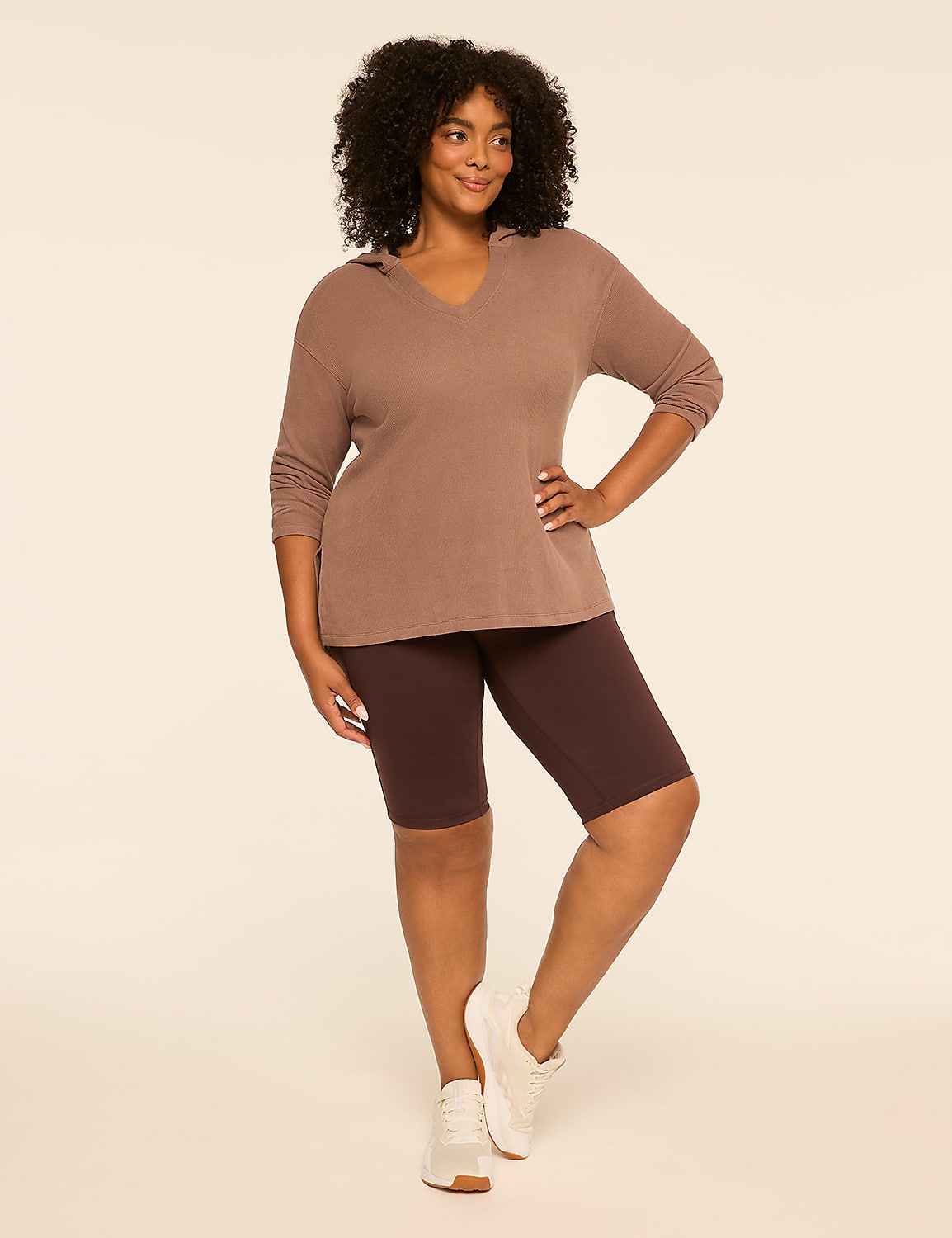 A woman in a brown hoodie and shorts stands against a plain, light brown background. Product Image 3