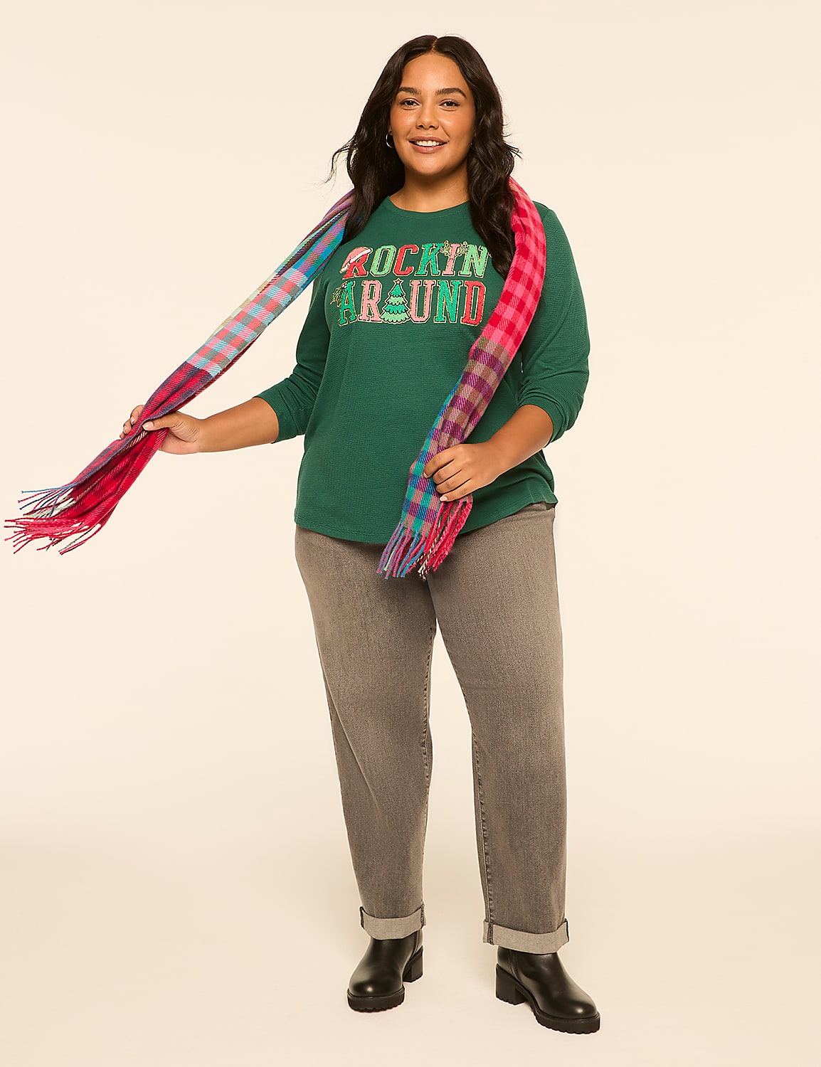 A smiling woman in a green "Rockin' Around" sweatshirt and gray jeans poses against a plain, light-colored background, with a colorful plaid scarf draped around her neck. Product Image 3