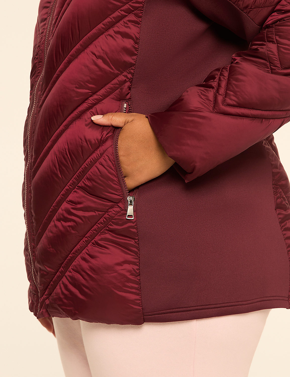A hand is placed in the zippered pocket of a burgundy quilted jacket with maroon side panels, against a plain cream background. Product Image 4