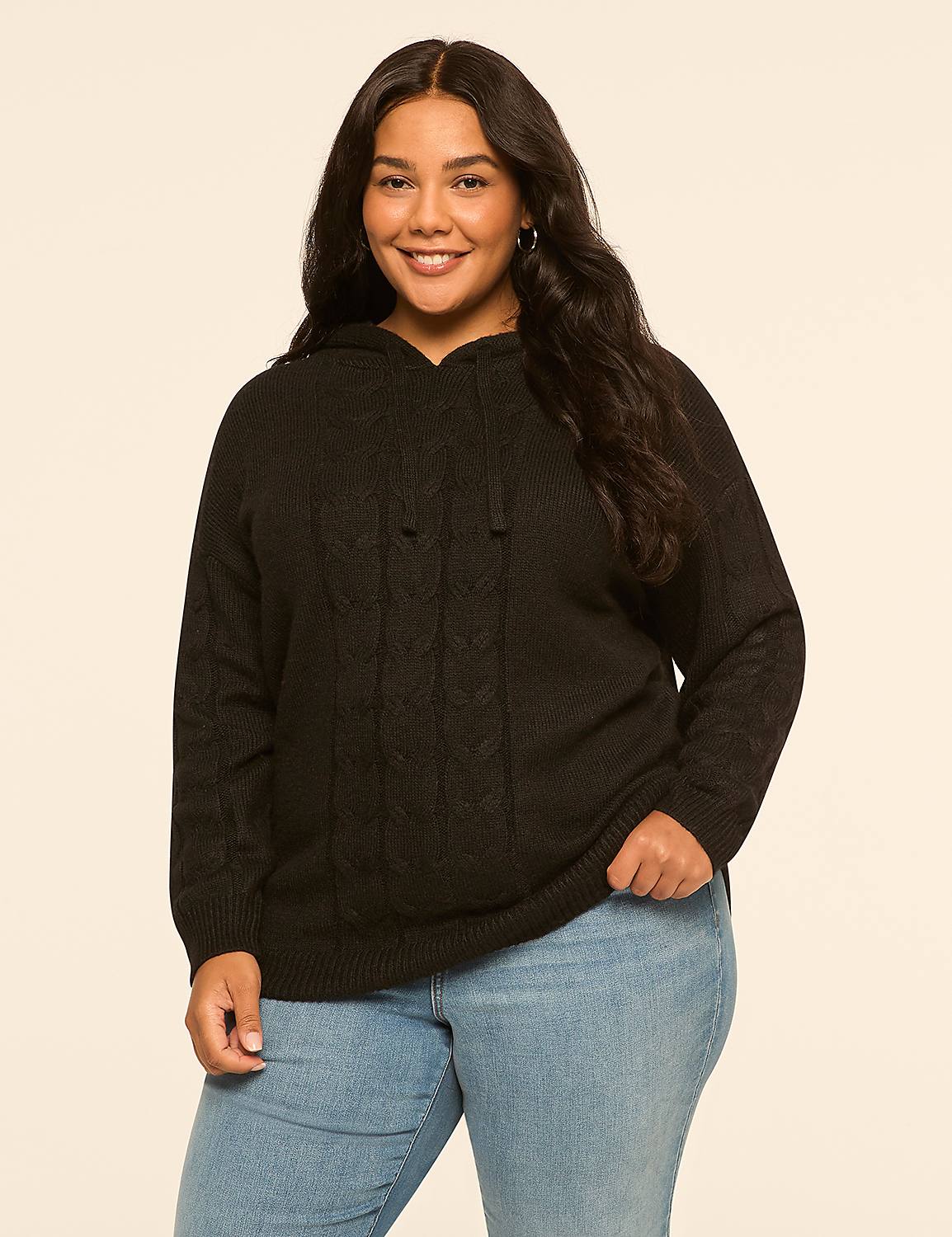 A smiling woman in a black cable-knit hoodie and light blue jeans stands against a plain, light beige background. Product Image 1