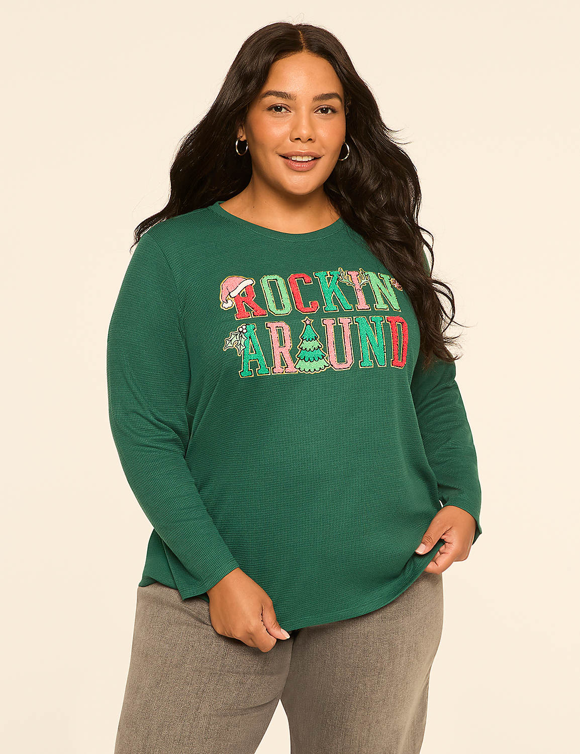 A woman in a green "Rockin' Around" Christmas-themed shirt and gray pants stands against a plain, light-colored background. Product Image 1