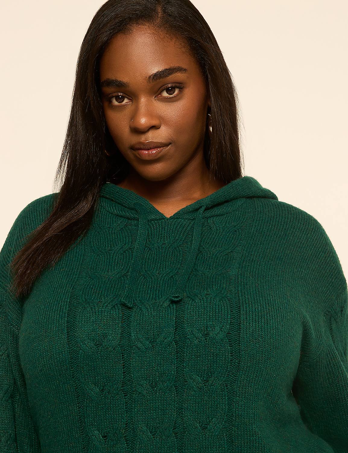 A woman with dark hair and brown eyes wears a green cable-knit hooded sweater against a plain, light-colored background. Product Image 4