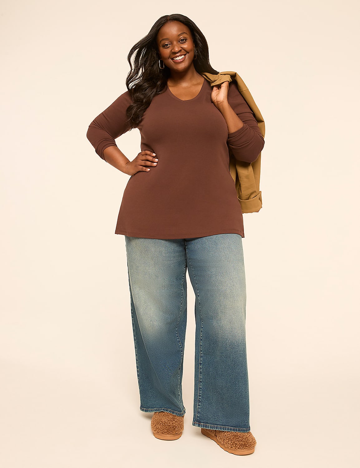 A smiling woman in a brown top and wide-leg jeans stands against a plain, light-colored background with a jacket slung over her shoulder. Product Image 3