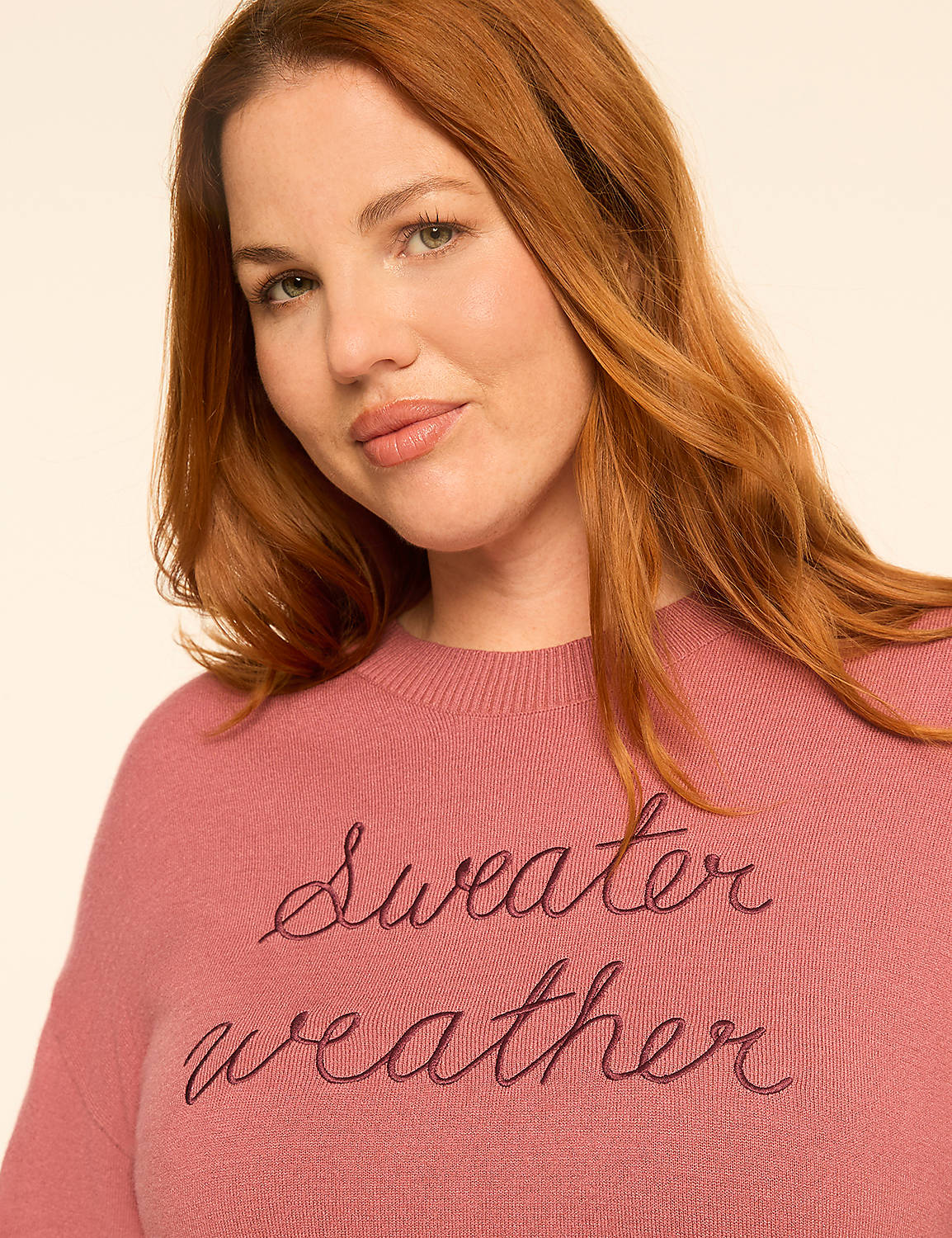 A smiling woman with curly hair wears a pink sweater with "Sweater weather" embroidered on it, against a plain background. Product Image 4