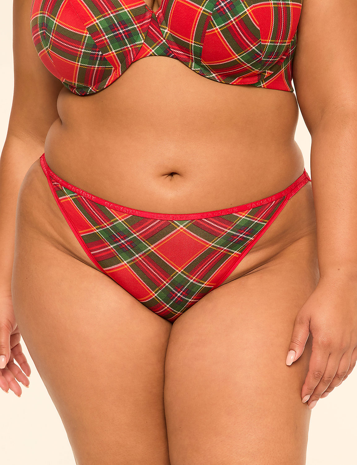A woman's torso is shown from the waist up, wearing a red, green, and yellow plaid bra and matching thong, against a plain, light-colored background. Product Image 1