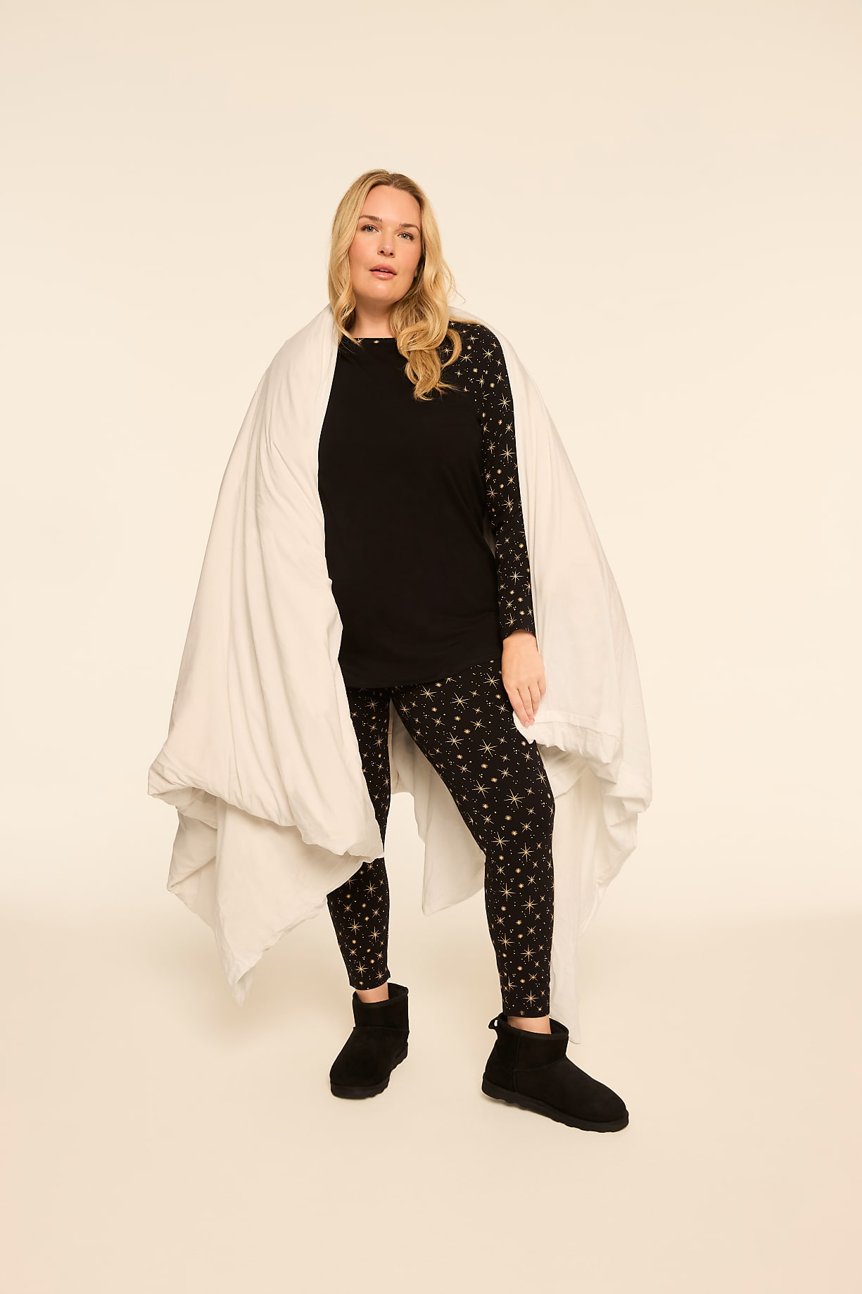 A woman stands in the foreground wrapped in a white duvet, wearing a black star-patterned pajama set and black slippers, against a plain, light-colored background. Product Image 6