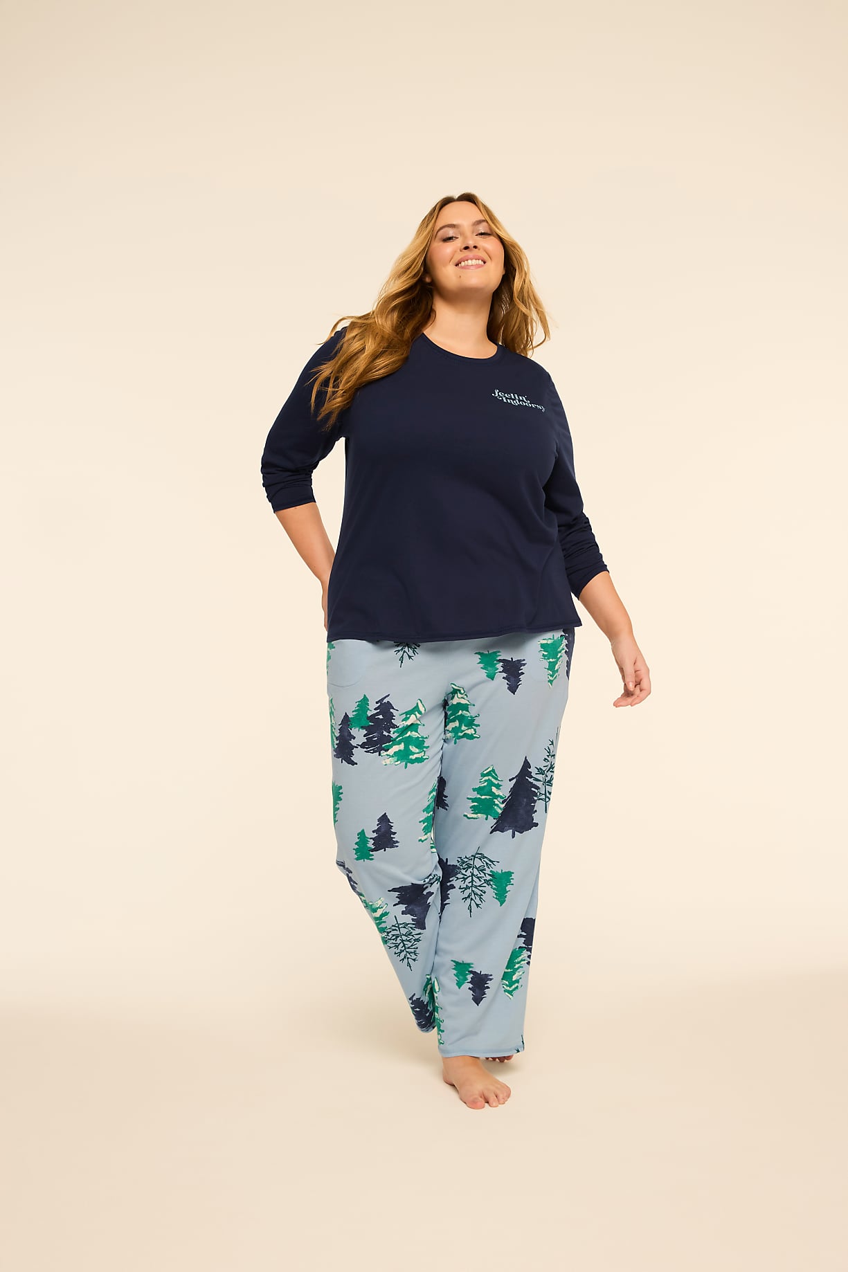 A smiling woman in a navy blue top and light blue pajama pants with a forest print stands against a plain, light-colored background. Product Image 5