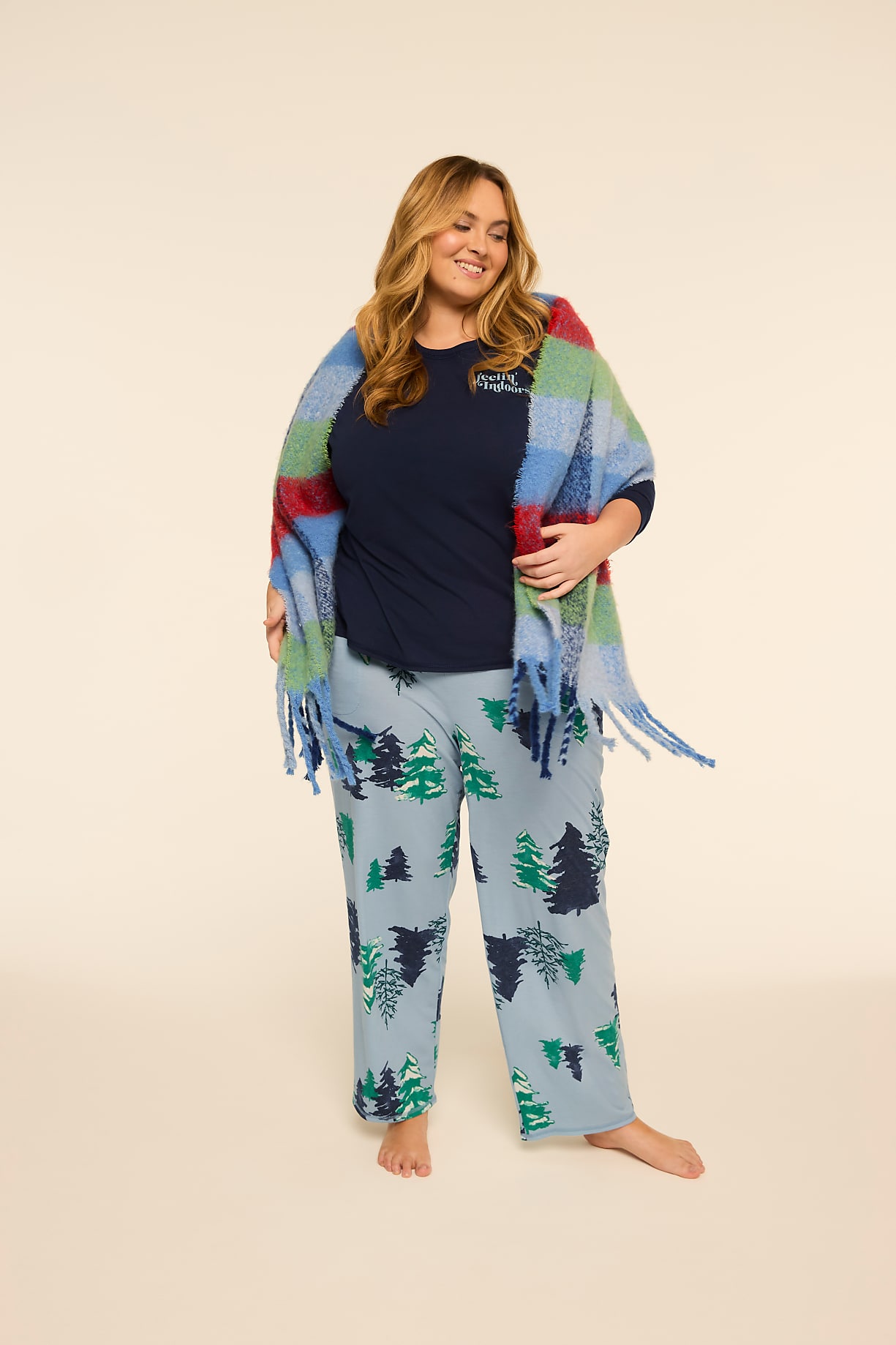 A woman in a navy shirt and light blue pajama pants with a forest print is wrapped in a colorful plaid scarf against a plain, light-colored background. Product Image 6