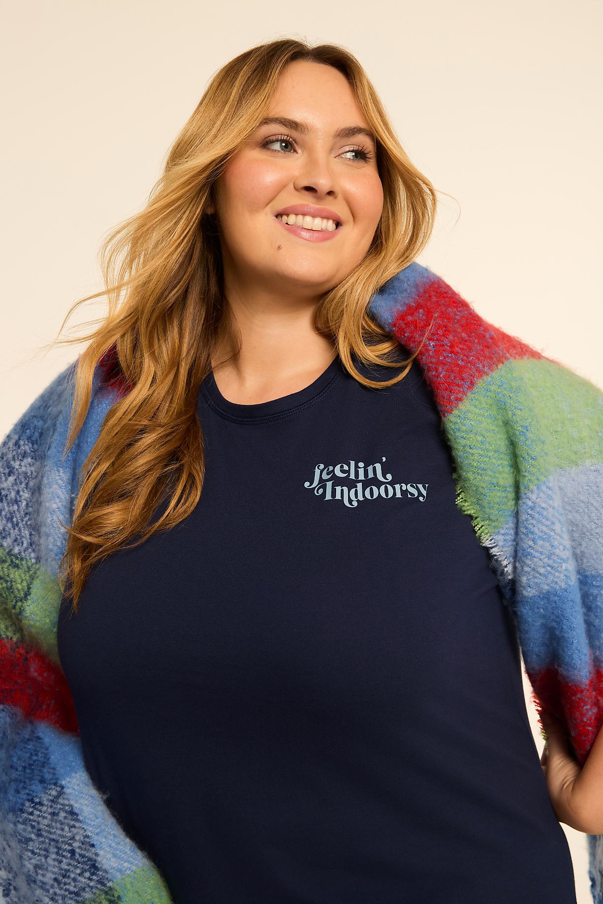 A smiling woman in a navy t-shirt with "feelin' indoorsy" printed on it is wrapped in a colorful plaid blanket against a plain, light-colored background. Product Image 9