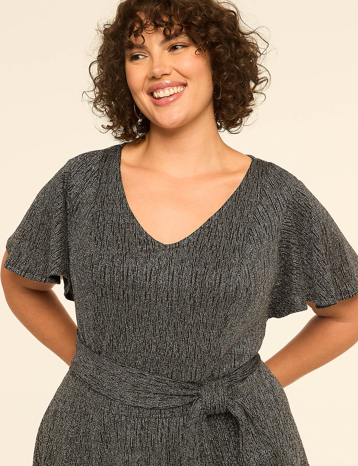 A smiling woman with curly hair wears a sparkly silver dress with flutter sleeves and a tie waist, set against a plain cream background. Product Image 3