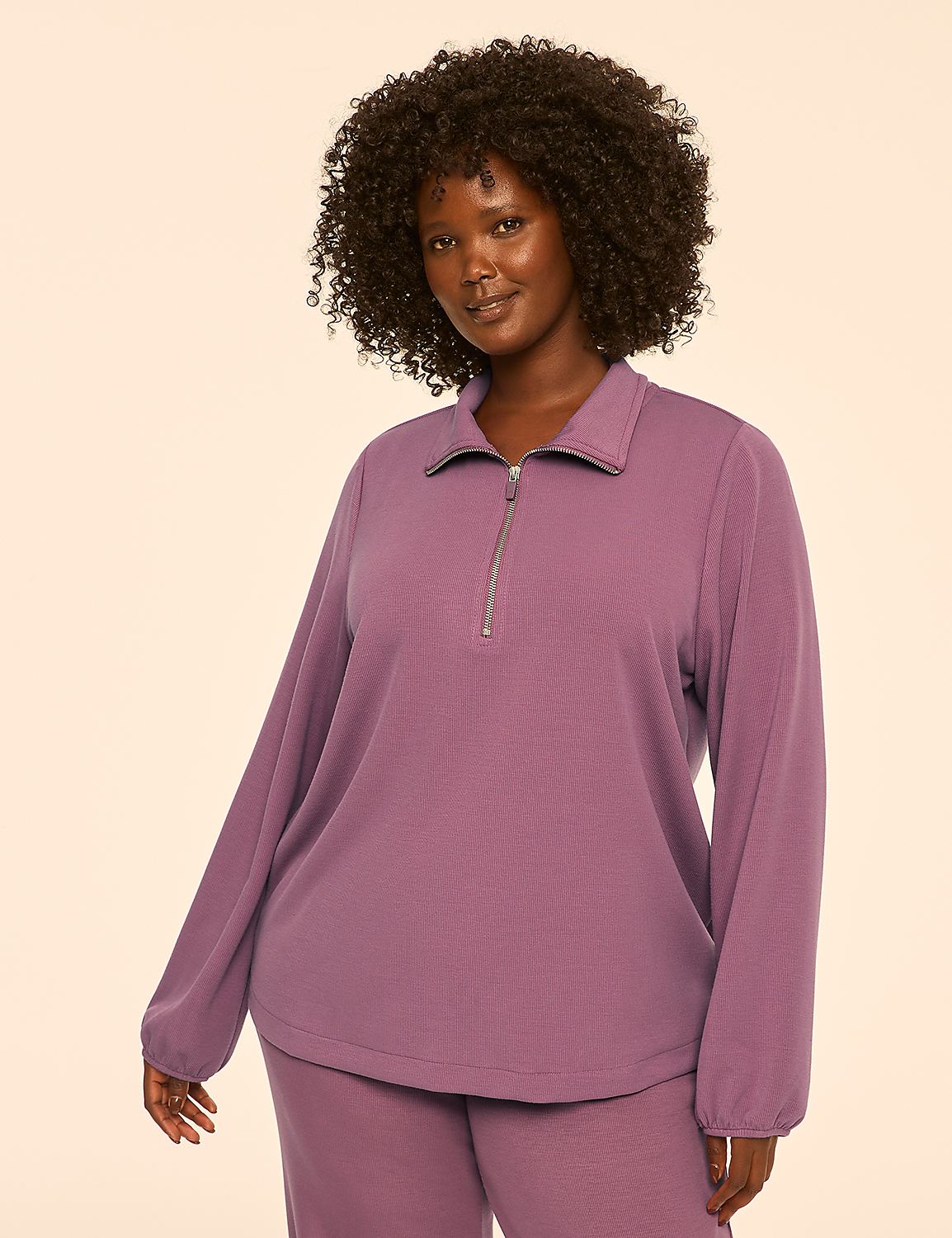 A woman with dark, curly hair and a warm smile poses in a mauve quarter-zip sweatshirt and matching pants against a plain, light peach background. Product Image 1