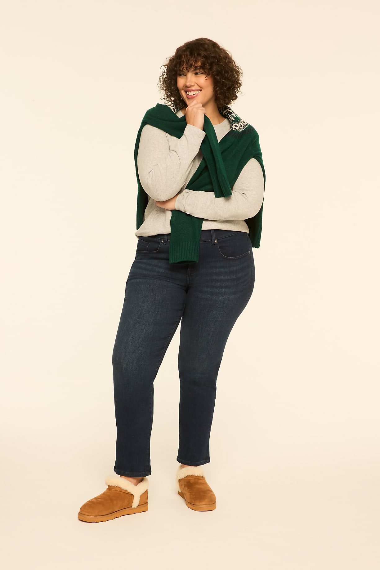 A woman wearing a light-colored top, dark blue jeans, and brown boots stands with a green sweater draped over her shoulders against a plain off-white background. Product Image 7