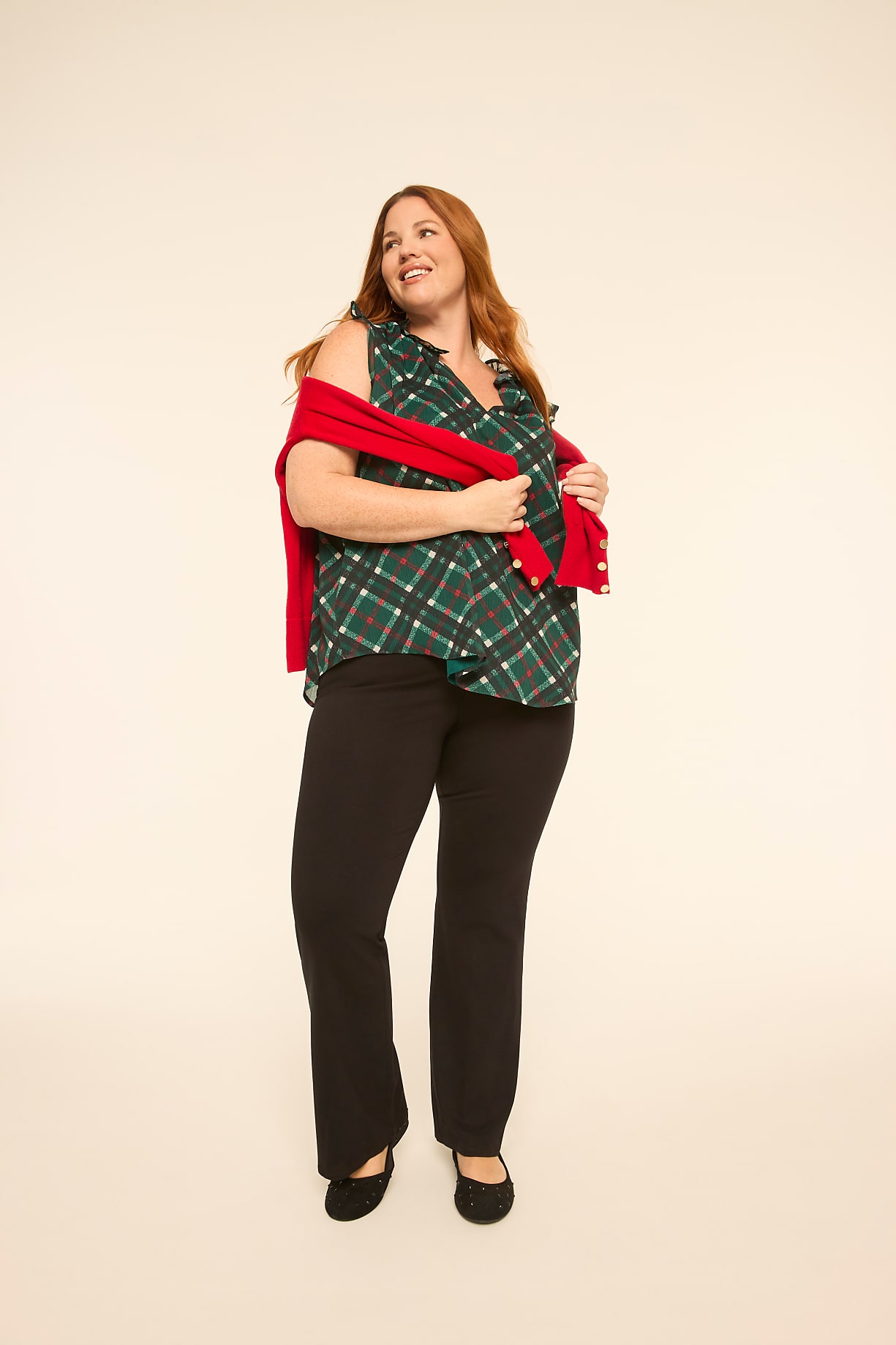 A woman wearing a green plaid shirt, black pants, and holding a red cardigan stands against a plain off-white background. Product Image 5