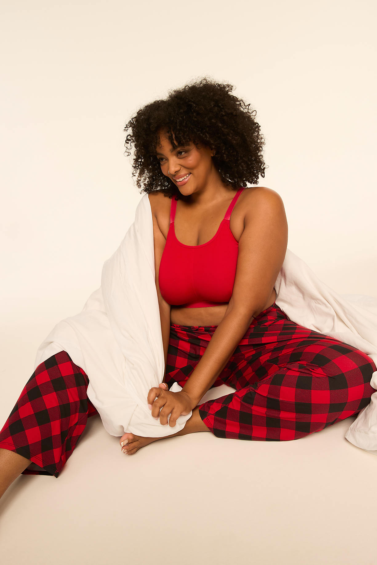 A smiling woman with curly hair wearing a red top and red-and-black plaid pajama pants sits wrapped in a white blanket against a plain, light-colored background. Product Image 7