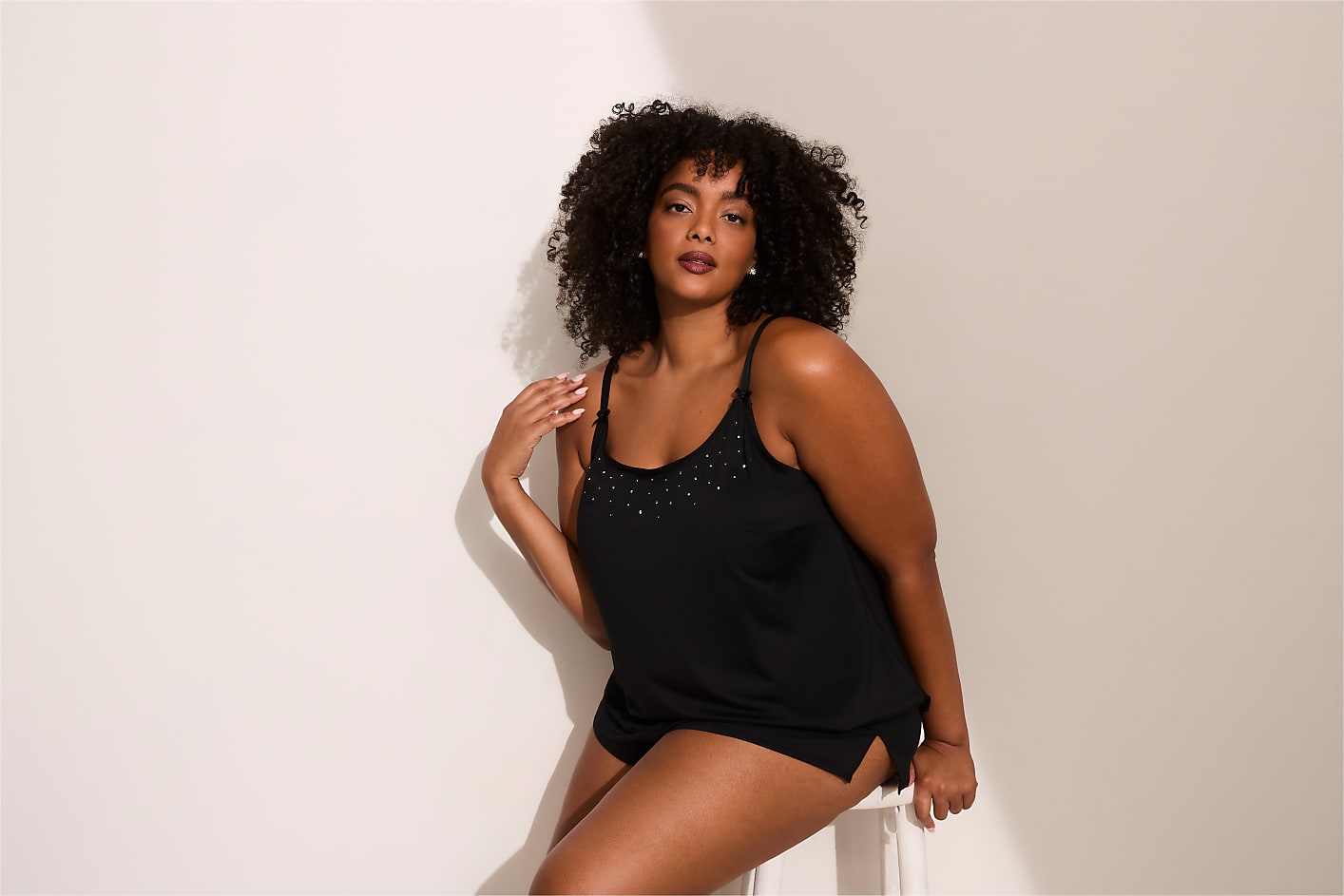 A woman with curly hair wearing a black slip dress poses against a plain white wall. Product Image 5