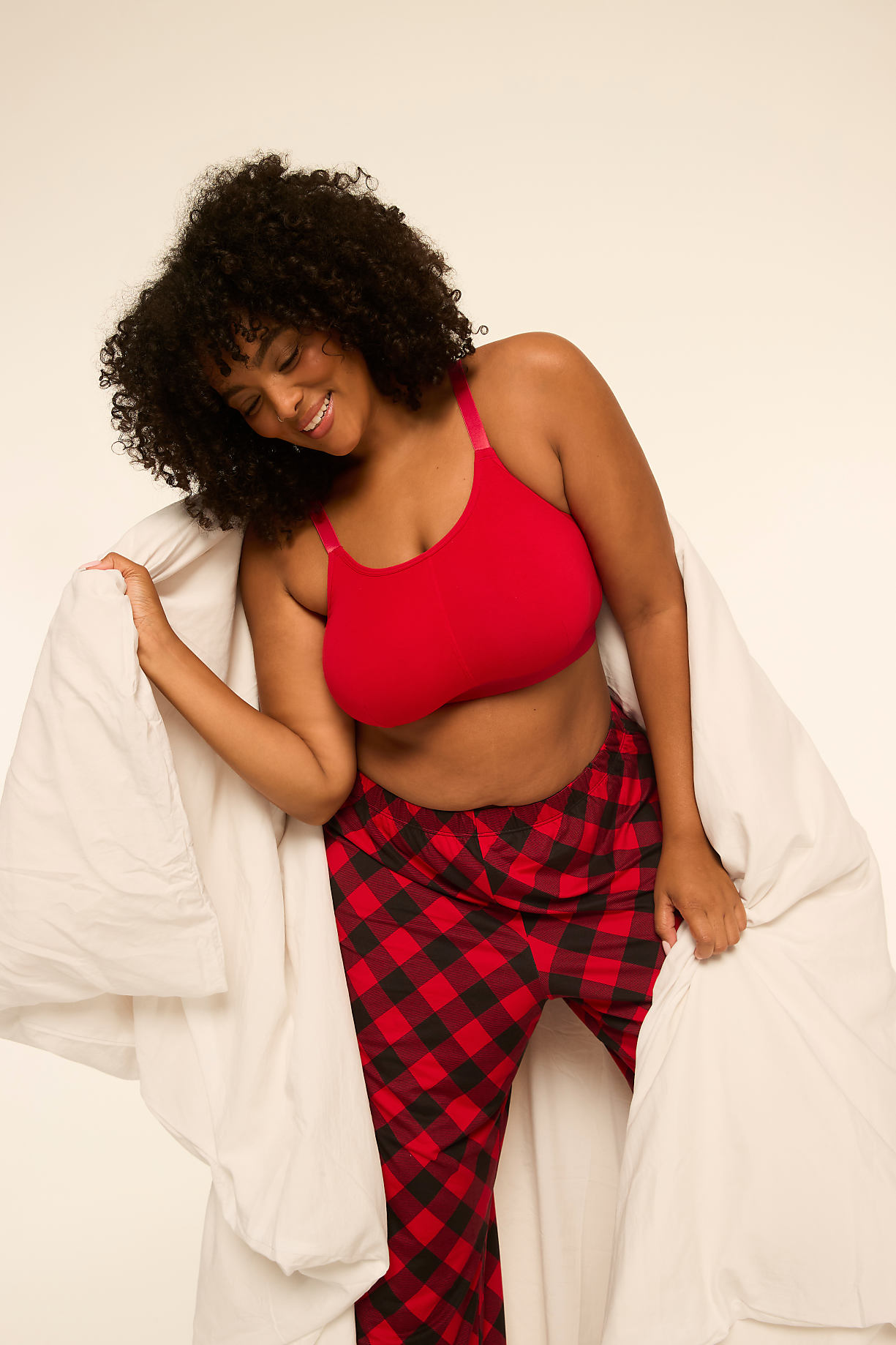 A smiling woman with curly hair wears a red bra and red-and-black checkered pajama pants while wrapped in a white duvet against a plain white background. Product Image 9