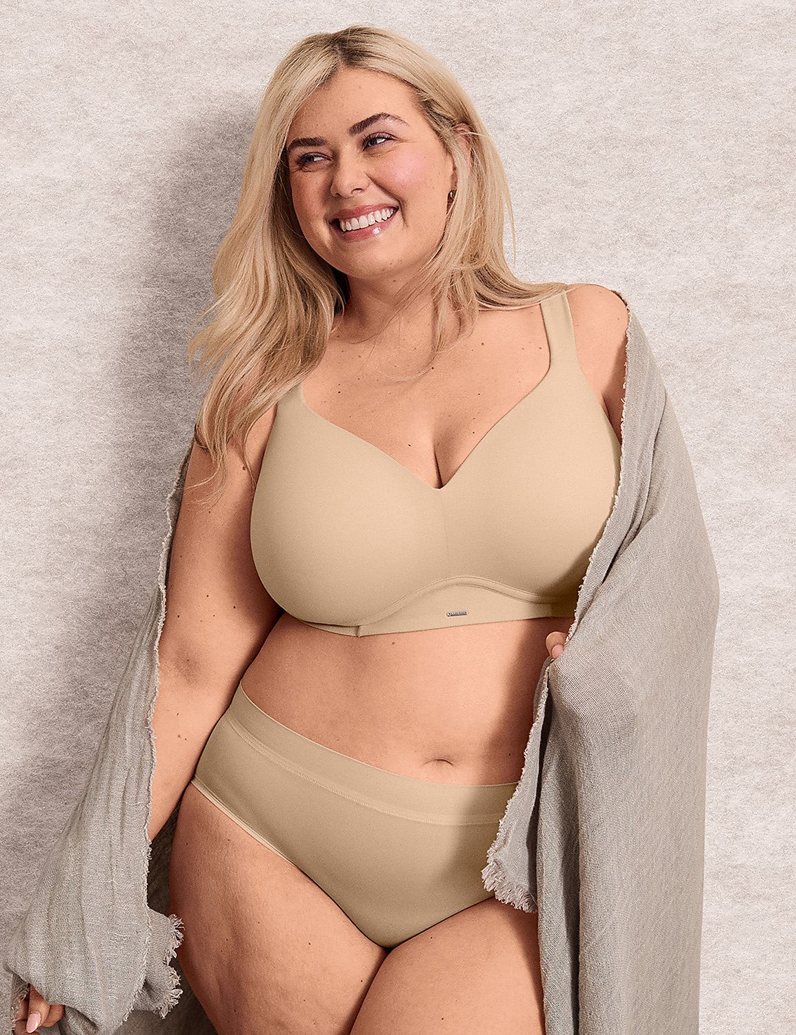 A smiling blonde woman in beige lingerie and a grey robe stands against a textured neutral wall. Product Image 5
