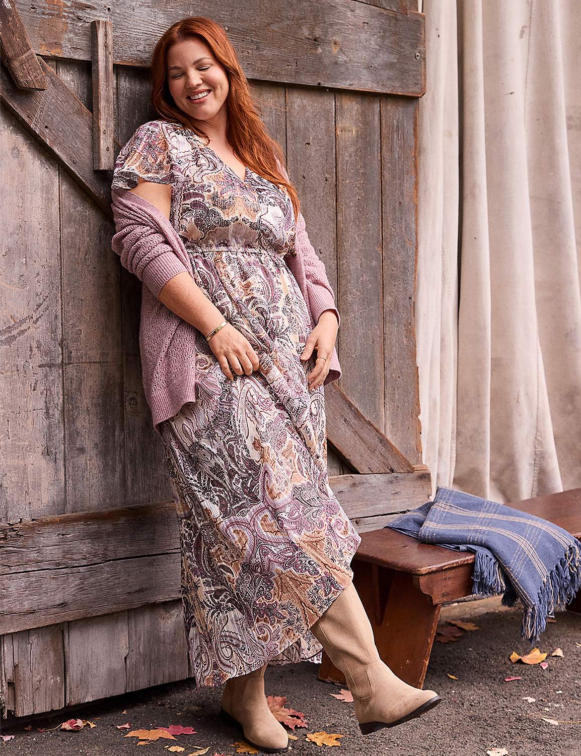 A smiling woman with red hair wearing a paisley dress, pink cardigan, and tan boots leans against a rustic wooden door next to a beige curtain and a wooden bench. Product Image 3