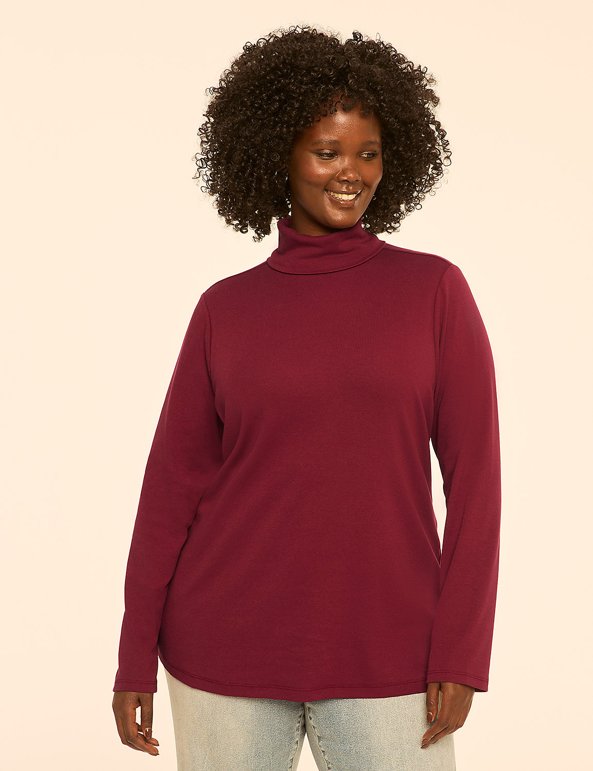 A smiling woman with curly dark hair wears a maroon turtleneck and light blue jeans against a plain cream background. Product Image 1