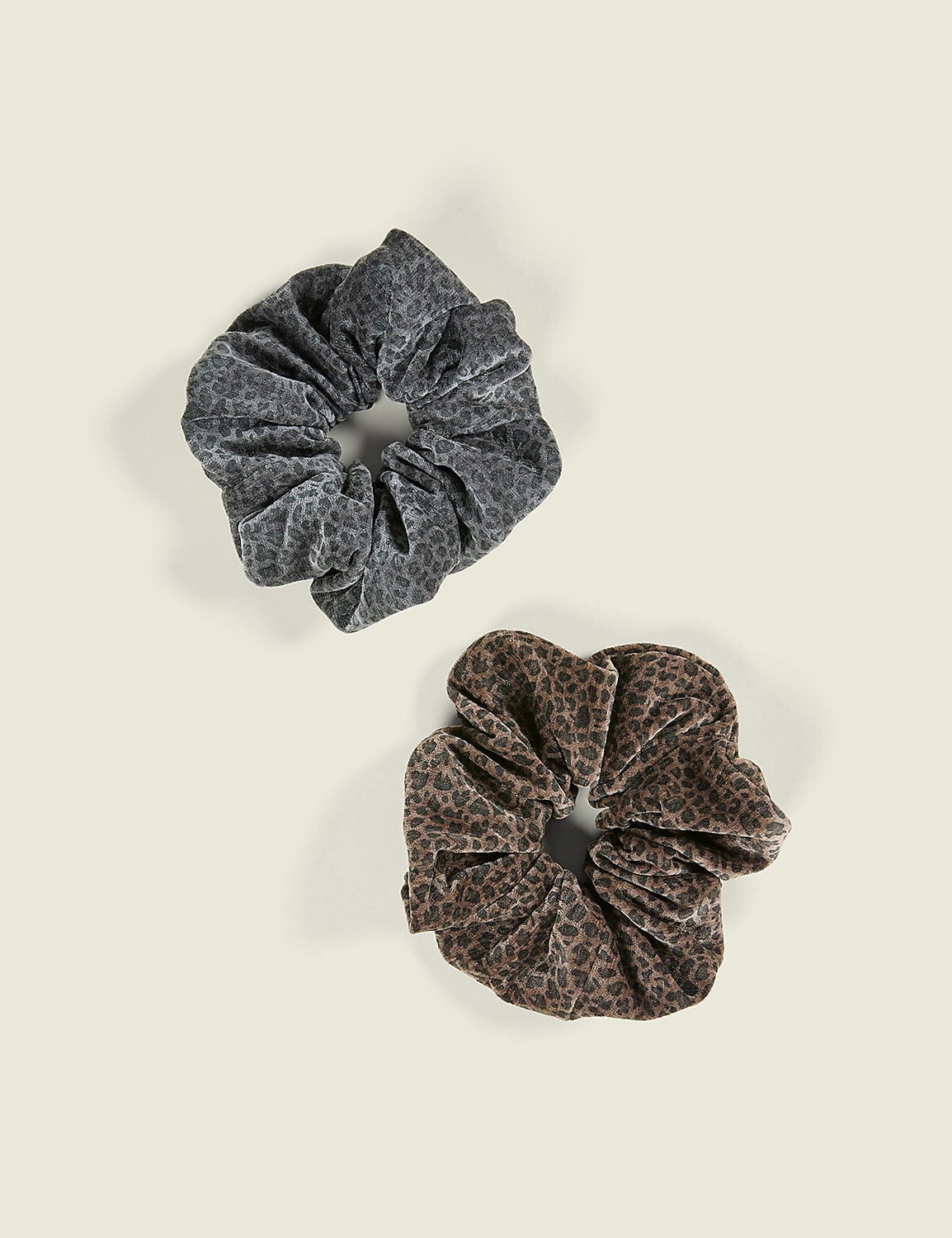 Two leopard-print scrunchies, one grey and one brown, lie on a plain off-white background. Product Image 1