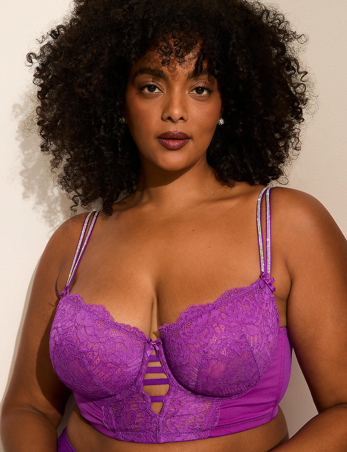 A woman with voluminous curly hair wearing a purple lace bra poses against a plain, light-colored wall. Product Image 2