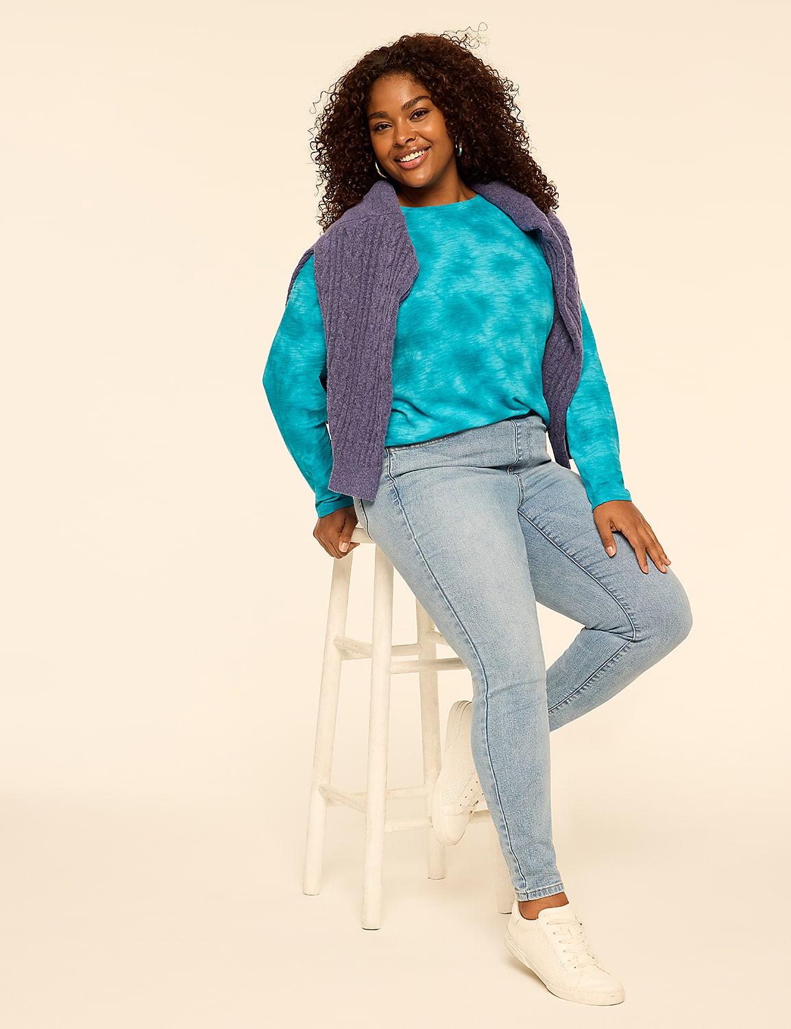 A smiling woman wearing a blue tie-dye shirt, purple vest, and light-wash jeans leans against a white wooden stool against a plain off-white background. Product Image 3