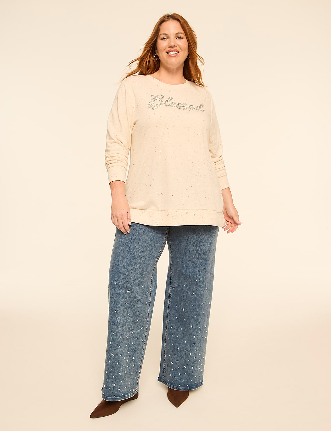 A smiling woman wearing a cream-colored "Blessed" sweater and wide-leg speckled jeans stands against a plain, light-colored background. Product Image 3