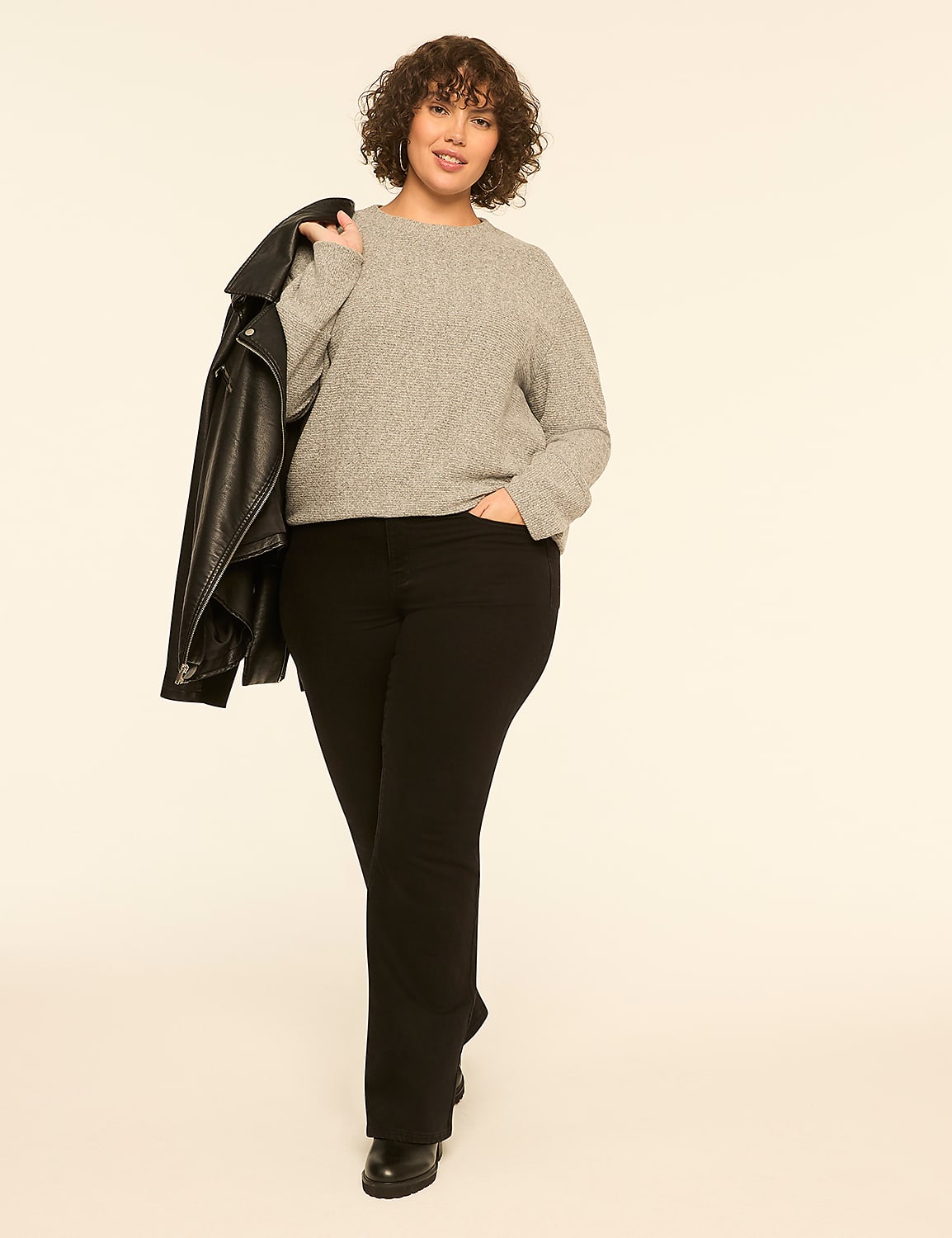A woman wearing a grey sweater and black pants holds a leather jacket over her shoulder against a plain white background. Product Image 3