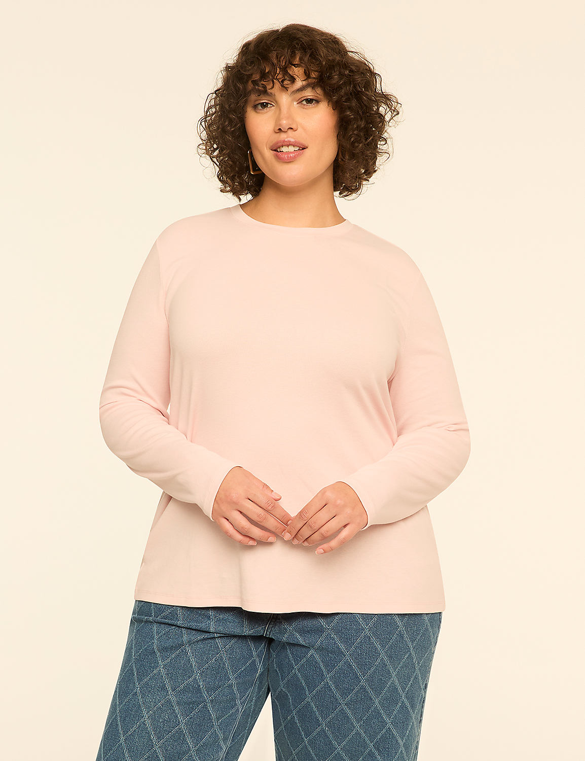 A woman with curly brown hair wearing a light pink long-sleeved top and patterned blue jeans stands against a plain off-white background. Product Image 1