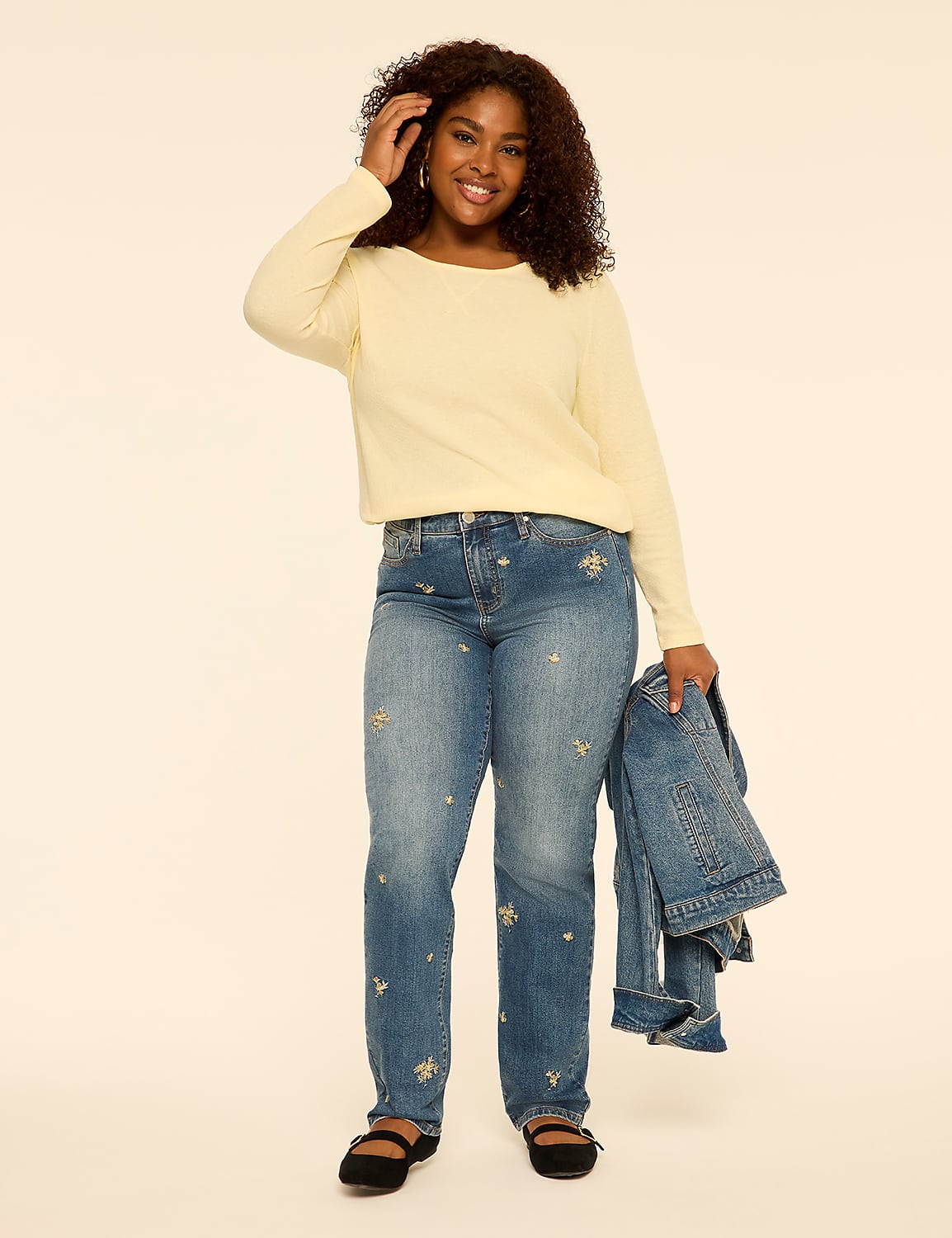 A smiling woman with curly hair wears a pale yellow long-sleeved top and embroidered blue jeans, holding a denim jacket against a plain, light-colored background. Product Image 3