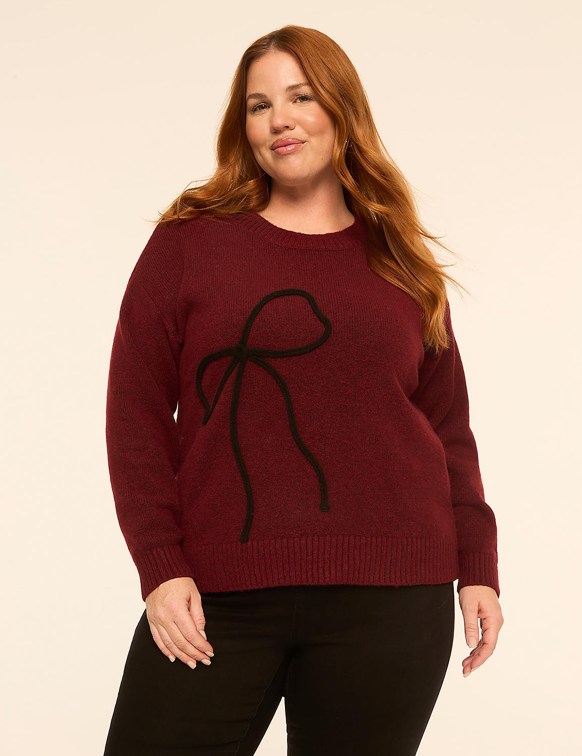 A woman with long red hair wearing a burgundy sweater with a black bow design and black pants stands against a plain off-white background. Product Image 1