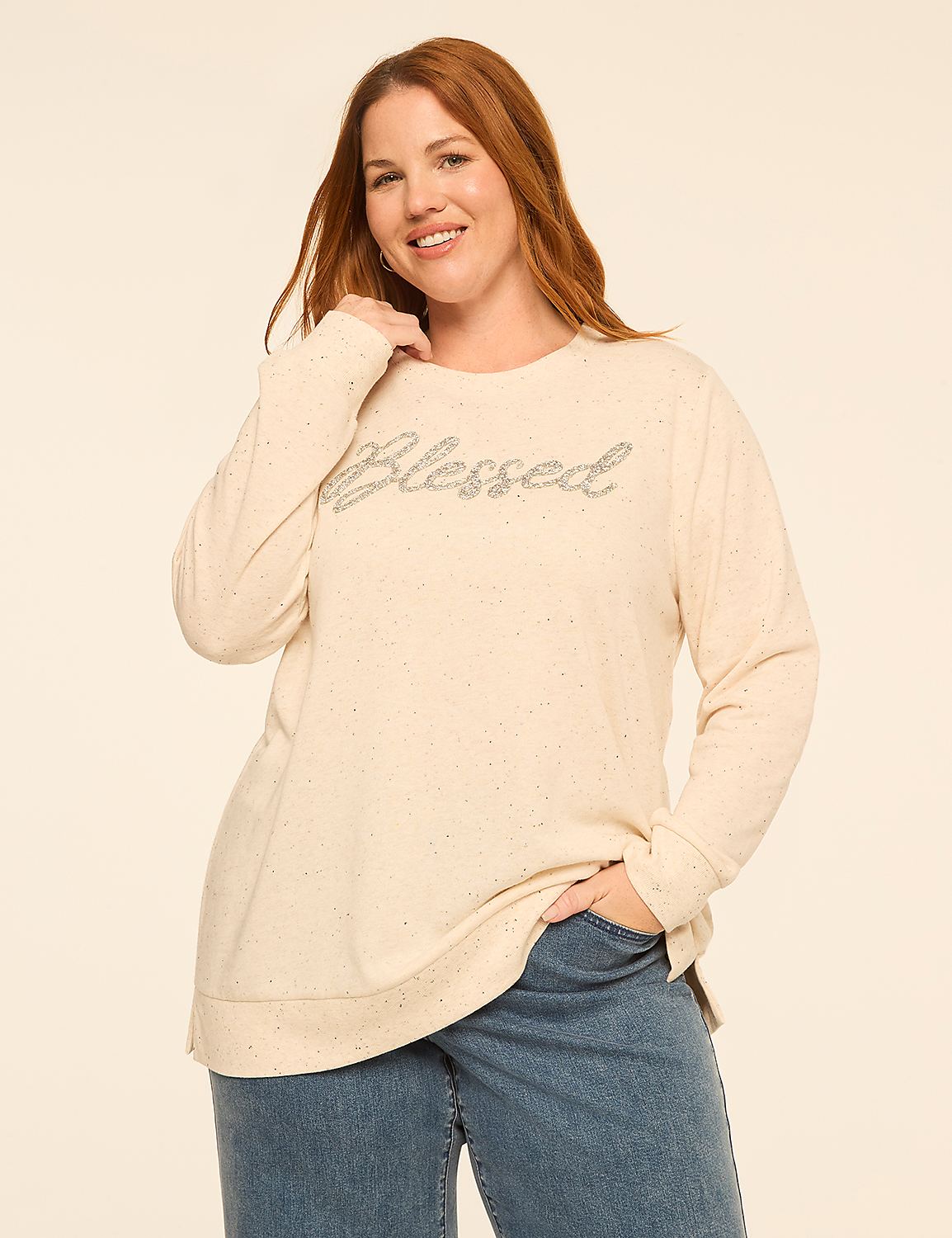 A smiling woman wearing a cream-colored "blessed" sweater and blue jeans stands against a plain, light-colored background. Product Image 1