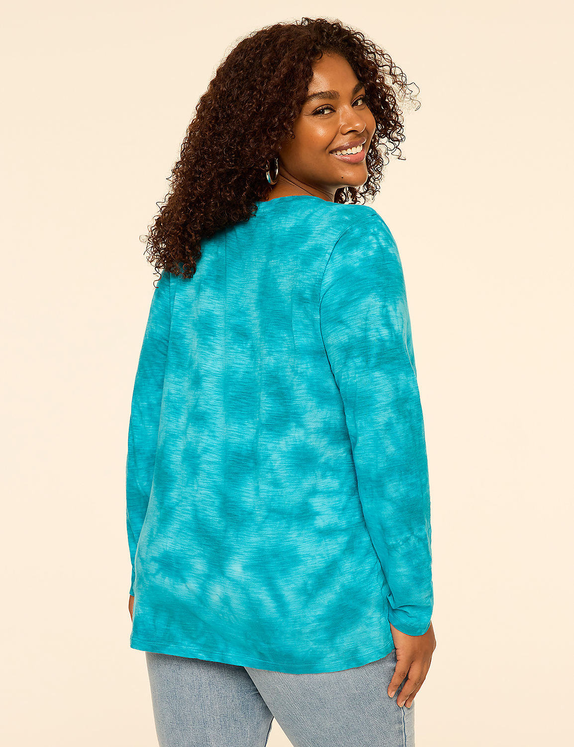A smiling woman with curly hair wears a blue tie-dye long-sleeve shirt and light-wash jeans against a plain, light-beige background. Product Image 2