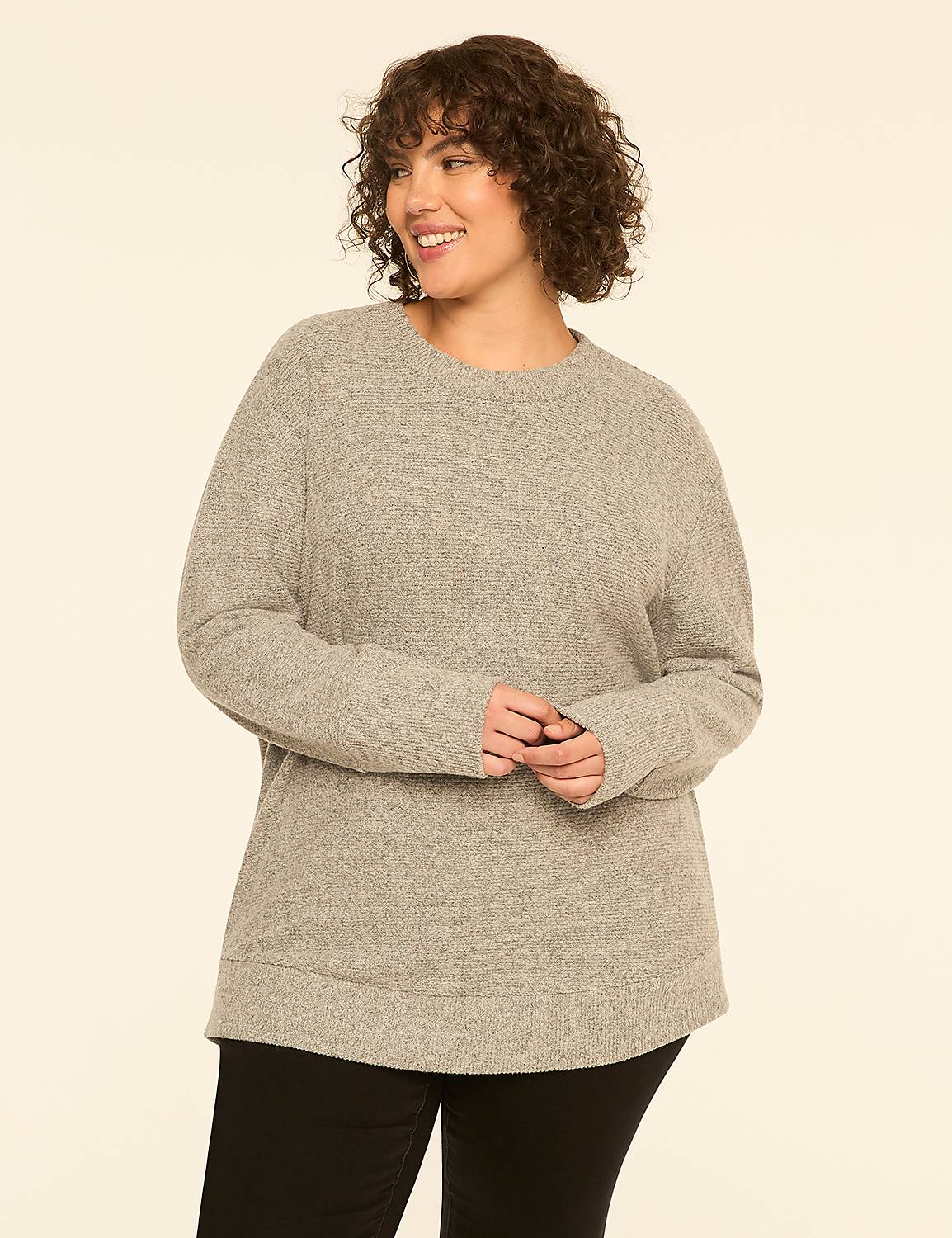 A smiling woman with curly brown hair wearing a grey sweater and black pants stands against a plain off-white background. Product Image 1