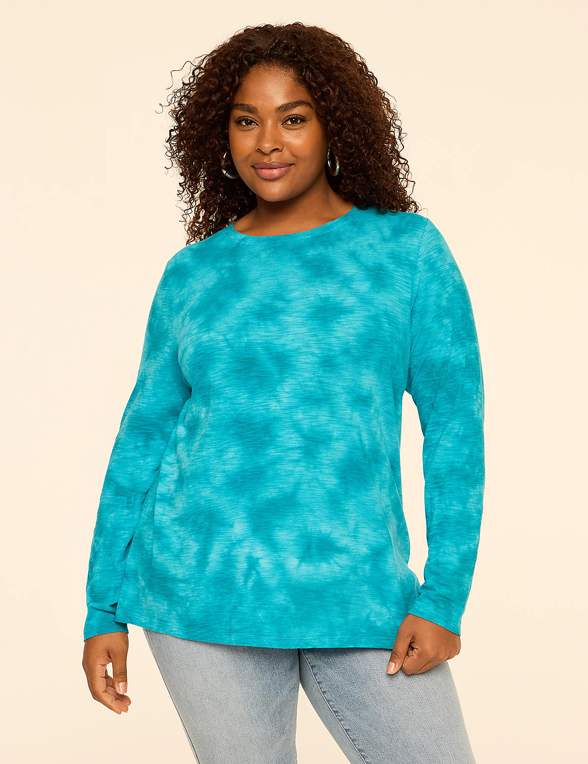 A woman with curly hair wearing a blue tie-dye long-sleeve shirt and light-wash jeans stands against a plain, light-beige background. Product Image 1