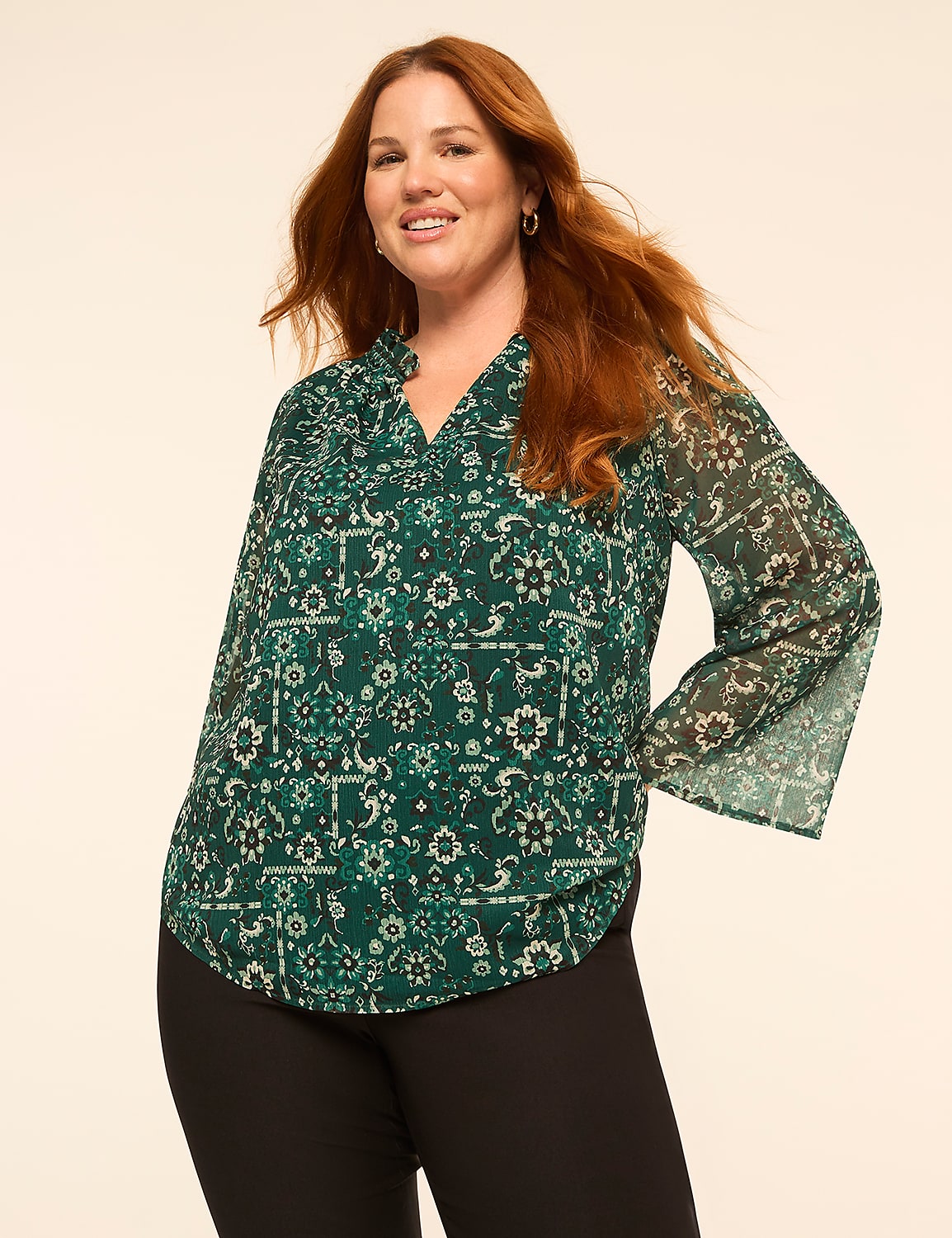 A woman with long red hair wearing a green patterned blouse and black pants stands against a plain off-white background. Product Image 1