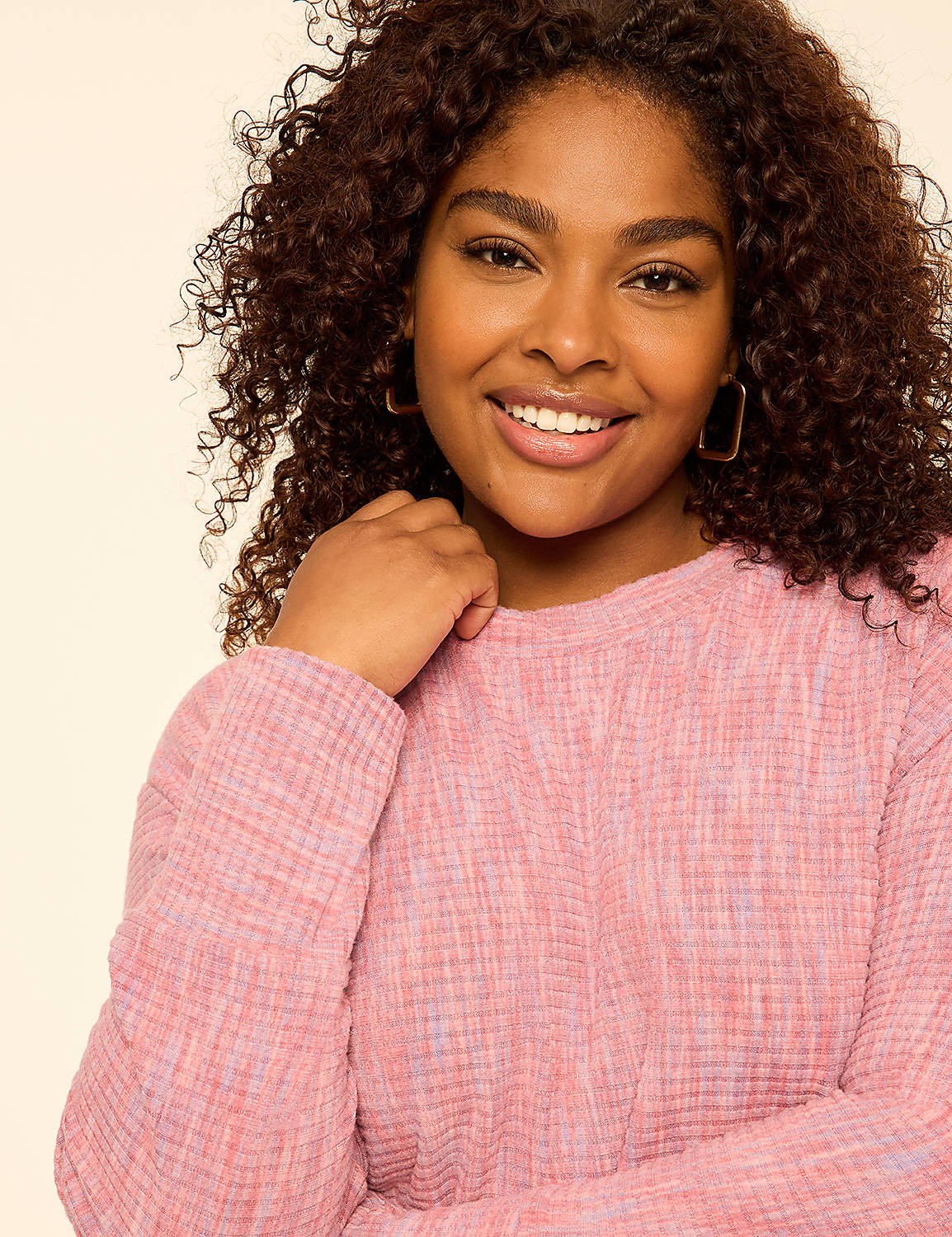 A smiling woman with curly brown hair wearing a pink textured sweater stands against a plain, light-colored background. Product Image 4