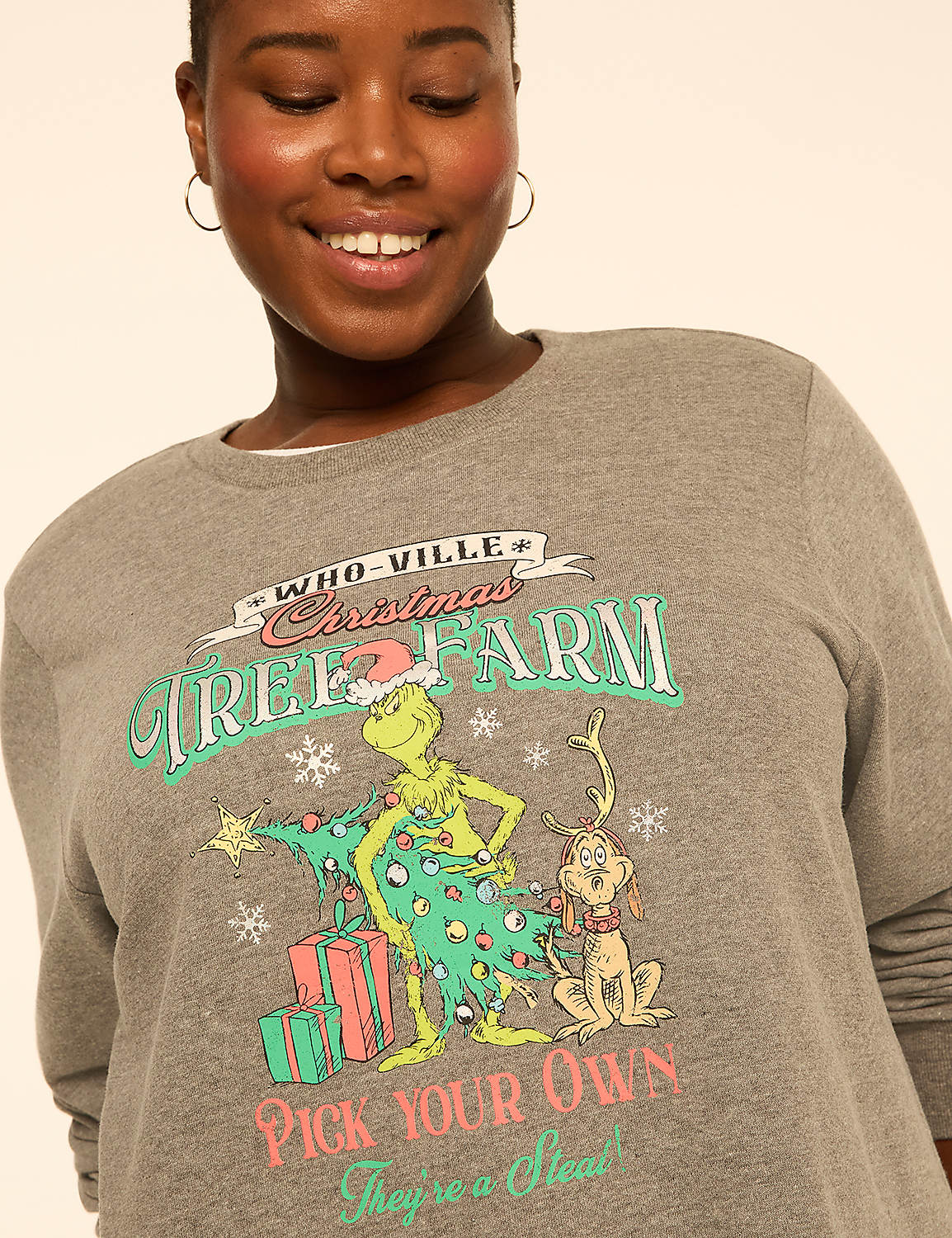 A smiling woman wears a grey sweatshirt featuring a Grinch-themed Christmas tree farm graphic against a plain, light-colored background. Product Image 4