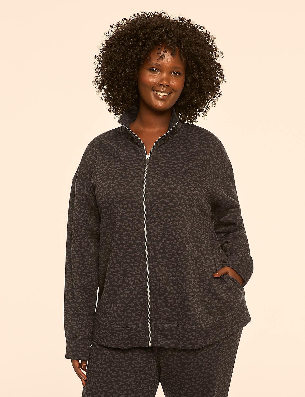 A smiling woman with curly dark hair stands in the foreground wearing a leopard-print zip-up jacket and matching pants against a plain, light-colored background. Product Image 1