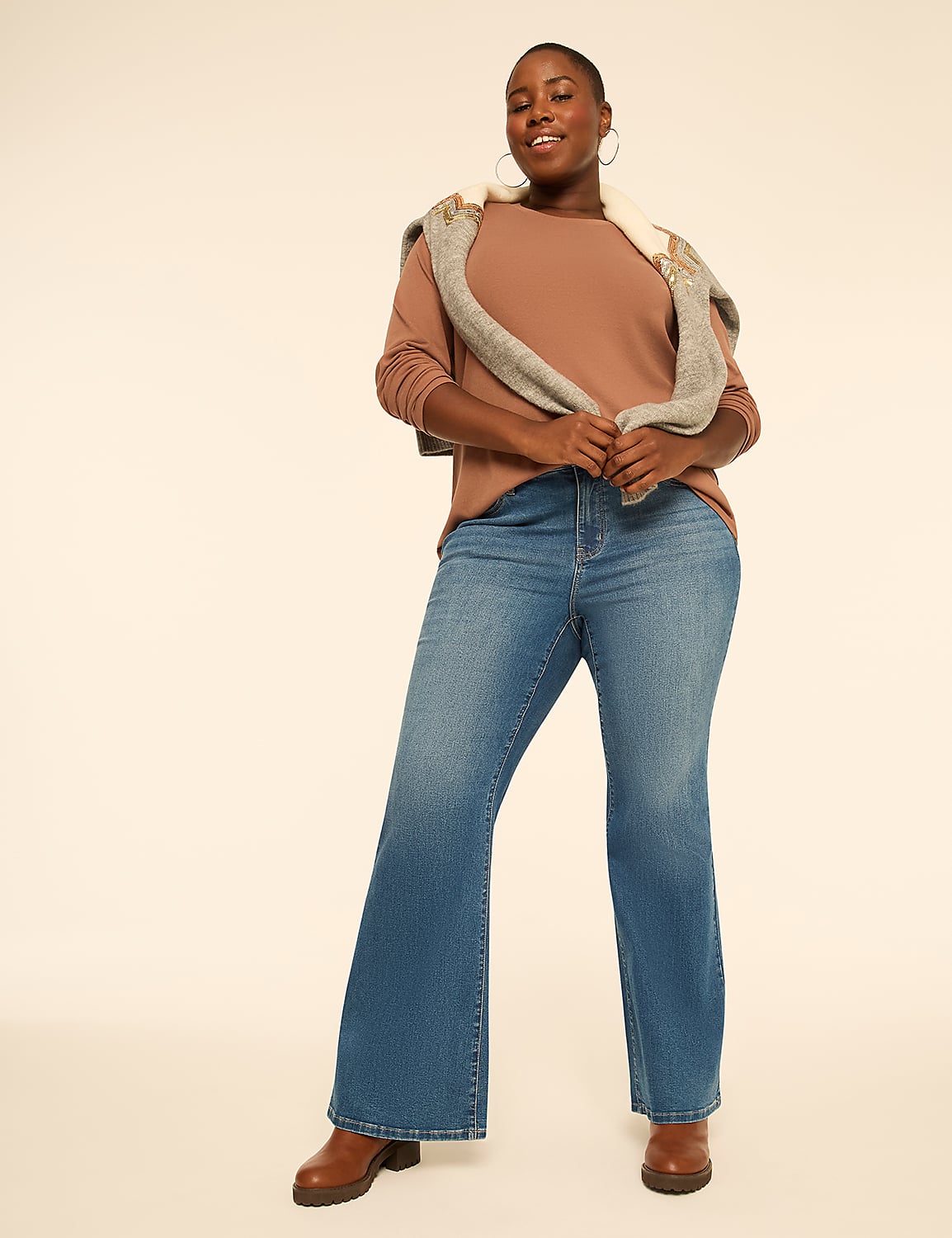 A smiling woman wearing a brown top, blue flared jeans, and a grey sweater draped over her shoulders stands against a plain, light-colored background. Product Image 3