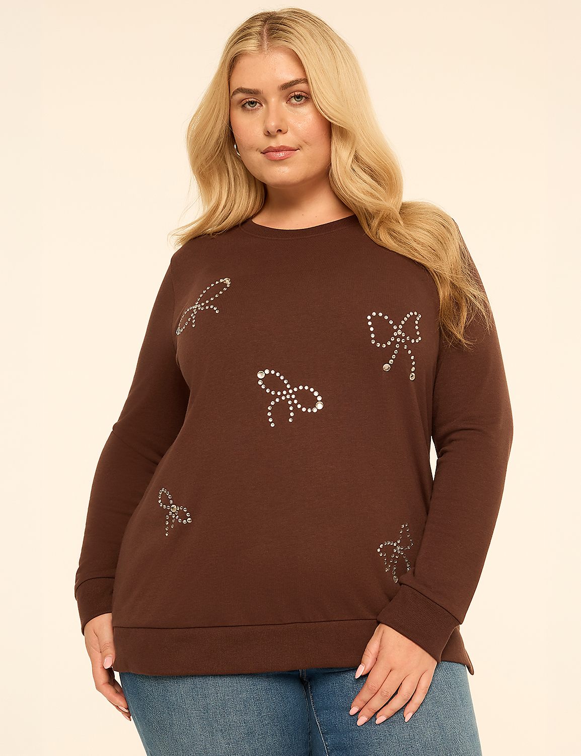 A blonde woman wearing a brown sweatshirt with rhinestone bows and blue jeans stands against a plain, light-colored background. Product Image 1