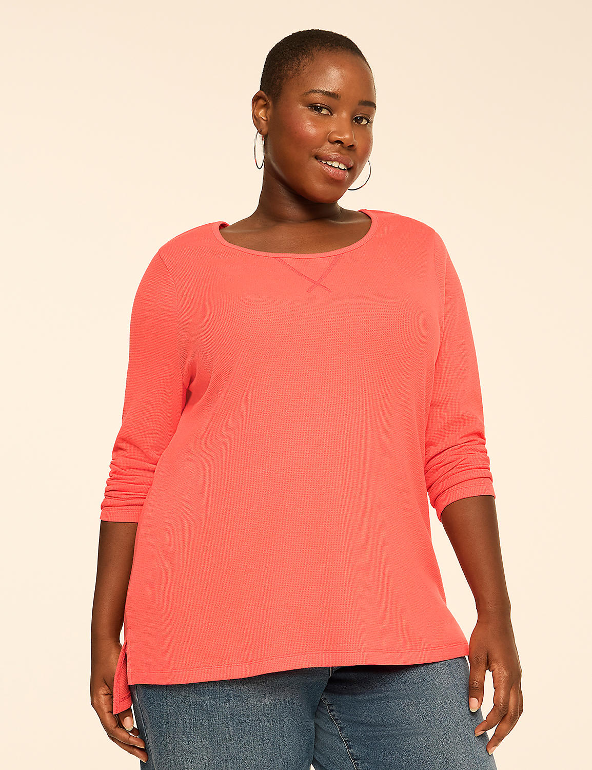 A woman wearing a coral-colored long-sleeved shirt and blue jeans stands against a plain, light beige background. Product Image 1