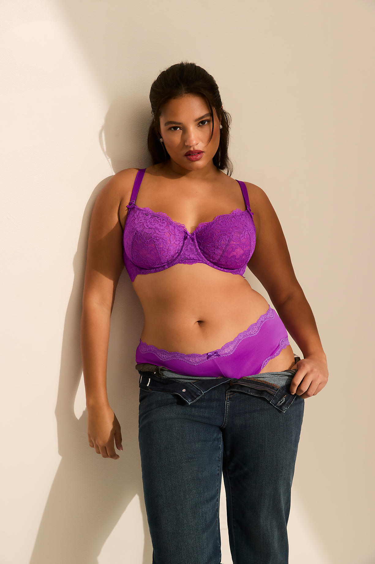 A woman wearing purple lace lingerie and unbuttoned blue jeans stands against a plain, light-colored wall. Product Image 10